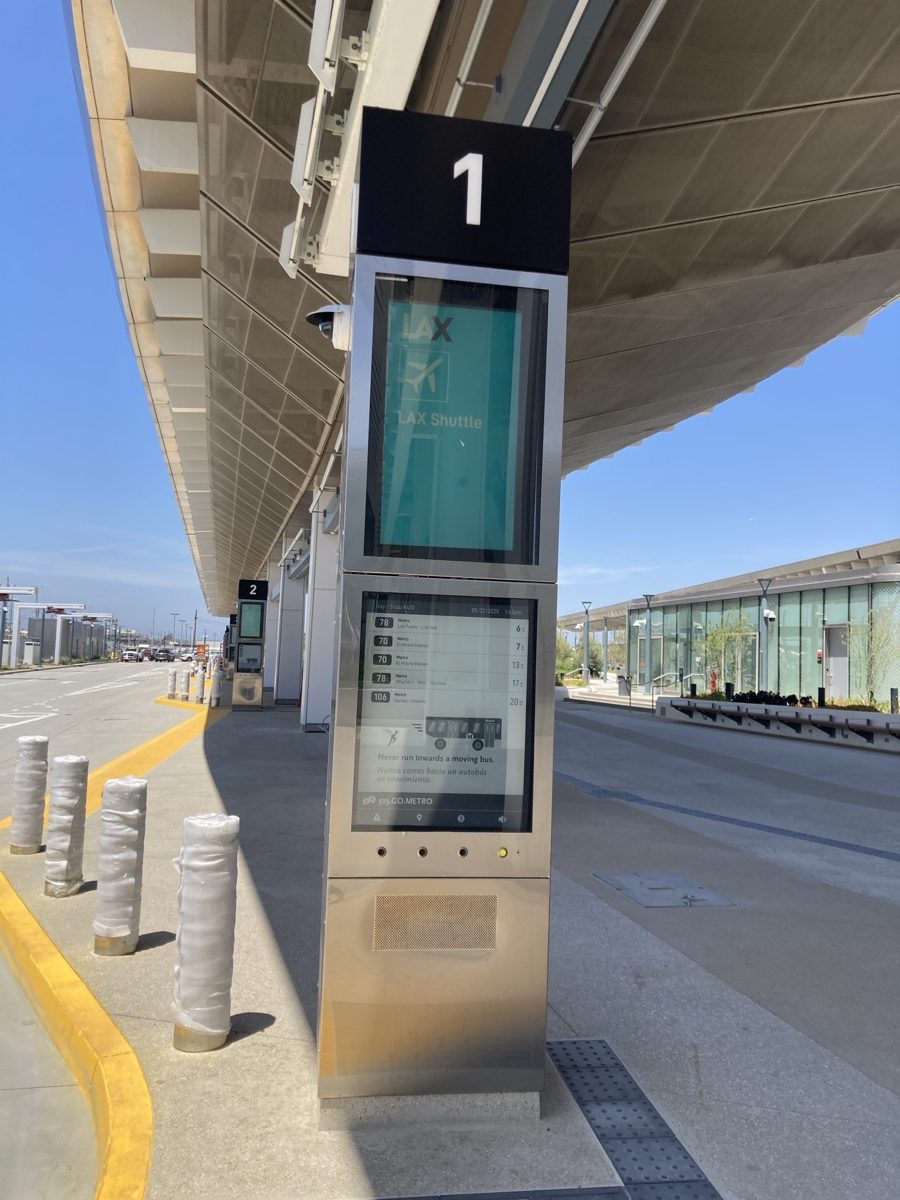 Stainless steel passenger information pylon at Bay 1 of the LAX/Metro Transit Center, with a digital screen showing LAX Shuttle above an e-paper screen listing upcoming Metro bus departures.