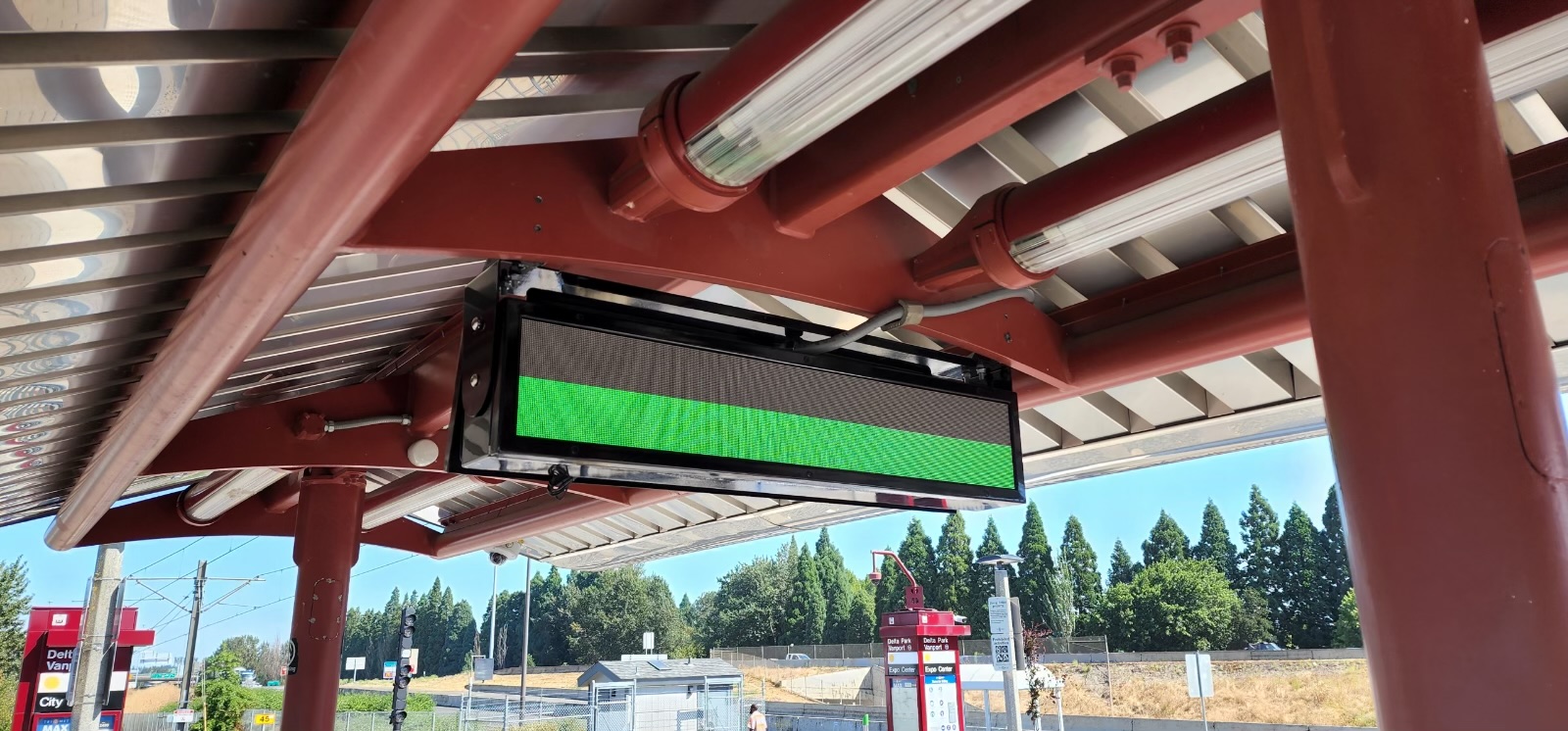 LED arrival sign mounted under the red steel canopy of the Delta Park/Vanport MAX platform in Portland, lit with a green test pattern during commissioning